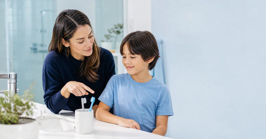 Parent and school-age child looking at two toothbrushes beside a bathroom sink.